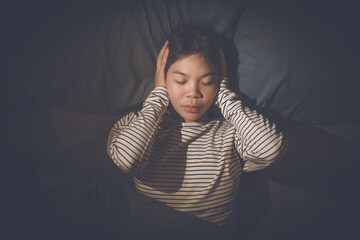 A young woman lies in bed, covering her ears with her hands, looking distressed, suggesting she's...