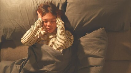A young woman lies in bed, covering her ears with her hands, looking distressed, suggesting she's trying to block out noise or suffering from a headache.