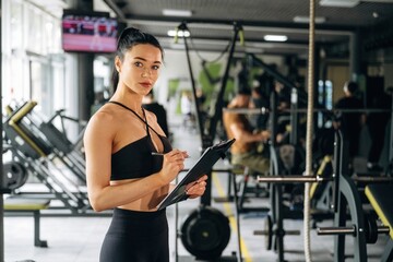 Fototapeta premium With notepad. Female trainer is standing in the gym