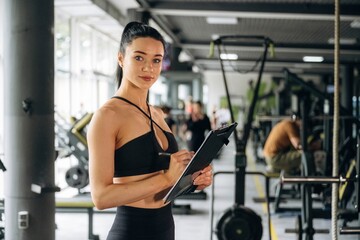 With notepad. Female trainer is standing in the gym
