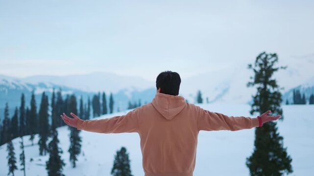 4K Rear view shot of an Indian male tourist raising his arms in front of the snowy Himalayan mountain peaks during the winter season at Gulmarg in Jammu and Kashmir, India. Man enjoys winter season.