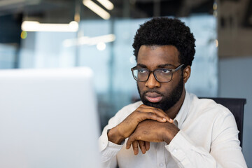A focused, man wearing glasses looks intently at a computer screen, deep in thought at work.