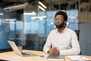 Obraz premium A thoughtful African-American man in glasses sits at a desk in an office, with a laptop and notepad.