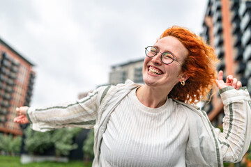 Joyful woman with curly orange hair enjoys a windy day in an urban park with modern buildings in...