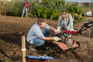 Male farmer shows his son how to fix garden cultivator. Farmer and a guy are repairing a machine to...