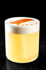 Close-up of a yellow cocktail with frothy white foam, orange peel garnish, and bitters drops in a clear glass on black background. Elegant alcohol drink perfect for bar or party themes.
