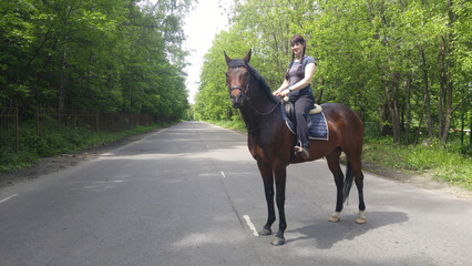 Woman is riding a brown horse on a asphalt road. The horse is standing on the road and the woman is...