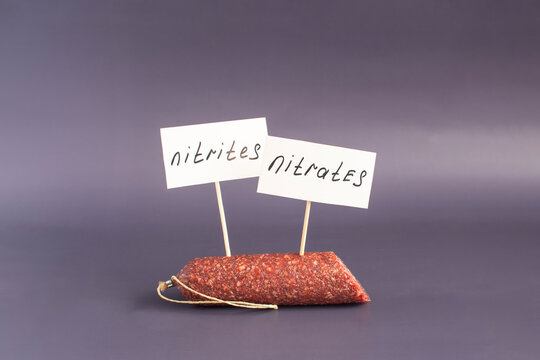 A fragment of smoked sausage and the warning labels "Nitrates and nitrites" in close-up on a dark background. The danger of harmful substances in sausage products