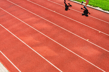 Illustrative athletics photo: runners’ legs and shadows on a red tartan track under clear daylight