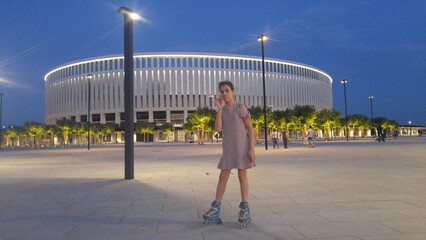 Girl is standing on roller skates in front of a large building stadium. She is wearing a black and...