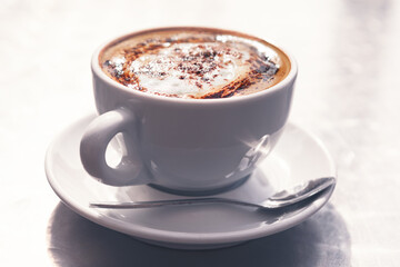 Creamy cappuccino in elegant white cup with saucer and spoon sits on sleek surface, topped with rich foam and cocoa. Good for coffee, breakfast or cozy lifestyle projects with warm, inviting appeal