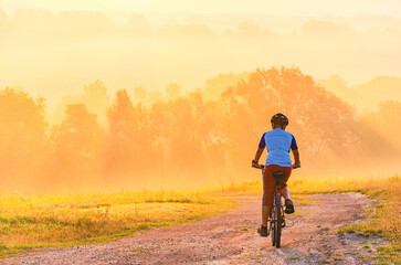 A man is riding a bike on a dirt road. The road is surrounded by trees. The sky is orange and the sun is setting