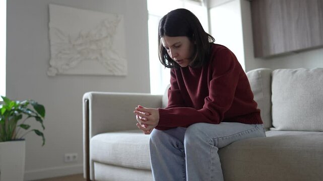Worried woman sits alone on sofa at home rubbing hands with nervous anxiety. Stressed female struggling with overwhelming thoughts, mental pressure, uncertainty. Distress, reflecting on life problems