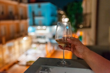 A woman's hand holds a glass of wine on the balcony ledge of a hotel in the summer evening, at night. Vacation time.