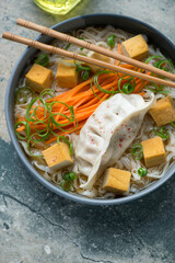 Bowl of rice noodles in miso broth with steamed dumpling, smoked tofu and vegetables, vertical shot, middle close-up, elevated view