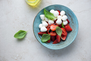Strawberry caprese with mini mozzarella and green basil in a turquoise bowl, horizontal shot on a light-beige stone background, top view