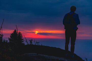 A man is standing on a hill with a backpack. The sky is orange and the sun is setting