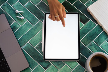Flat lay of a woman's hand interacting with a digital tablet screen, surrounded by a notebook, stylus, smartphone, and coffee on a vibrant green tiled surface. Ideal for digital work concepts.