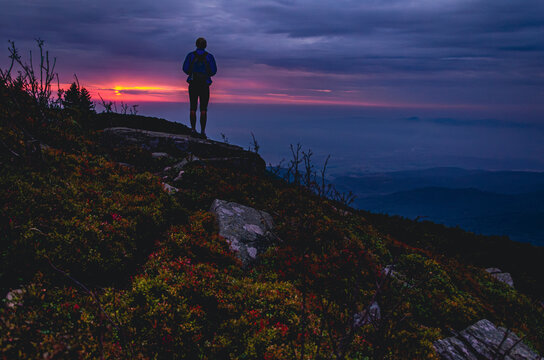 A man is standing on a hill with a backpack. The sky is orange and the sun is setting