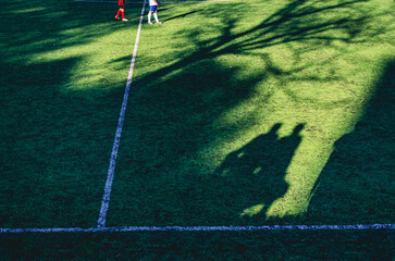 A soccer field with a shadow of a person on the grass. There are two people on the field