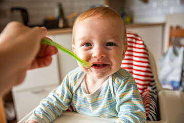 little cute boy 8 months old with blond hair and blue eyes eats porridge with a spoon in a high chair