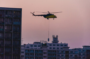 A helicopter is flying over a city. The helicopter is yellow and black. The sky is pink and purple