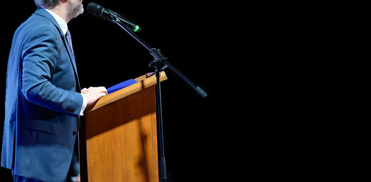 Side view of a man in a suit speaking into a microphone at a podium on a black background.