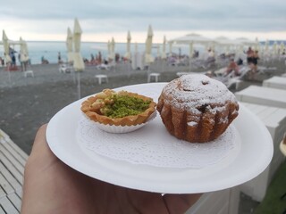 Person is holding a plate with a pastry and a muffin on beach sea with umbrellas. The pastry is...