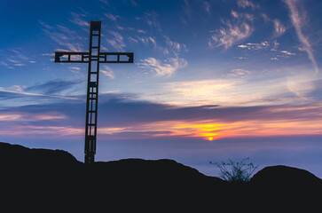 A cross is standing on a hillside with a beautiful sunset in the background. The sky is filled with...