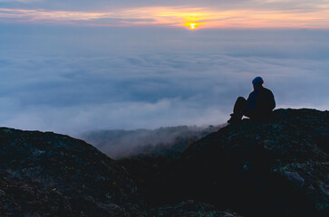 A man sits on a rock overlooking a foggy mountain. The sky is orange and the sun is setting