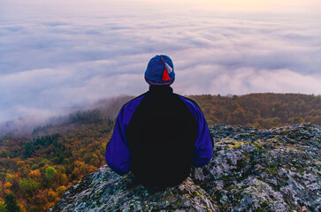 A man is sitting on a rock with a hat on. The sky is cloudy and the man is looking at the clouds