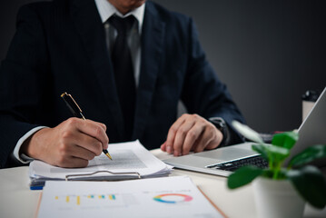 Hands holding pen signing paperwork on desk with laptop, representing contract signing, important decision-making, and corporate governance in a professional office.