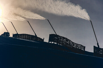 A group of snow guns are shooting water into the air, creating a misty atmosphere. The snow guns are positioned on a hill, possibly in a ski resort or a snow-covered area