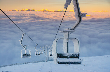 A ski lift with a chair covered in ice. The chair is located on the top of the lift, and it is surrounded by snow. The lift is located in a mountainous area, and the sky is cloudy