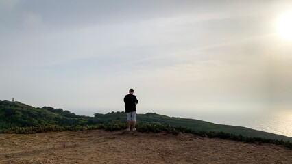 Solitary figure stands on a misty coastal cliff, contemplating horizons, reminiscent of Obon rituals or Earth Day