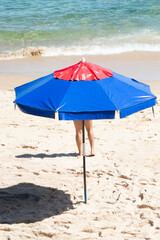 Unidentified person standing behind a red and blue umbrella on the seashore during strong sunshine.