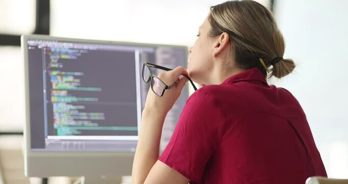 Female software developer works on new app program holding glasses in hand and looking thoughtfully at monitor. Focus on solving programming problem