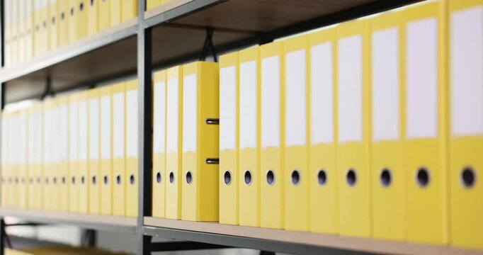 Office worker grabs red ring binder on shelf filled with yellow ring binders in archive. Woman focuses on organizing important documents and files