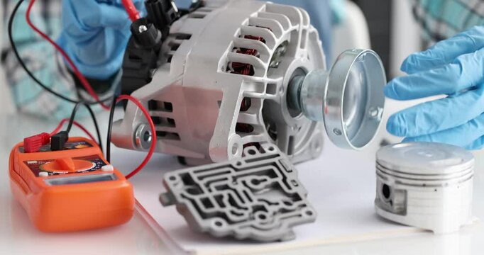 Gloved technician hands test car alternator with multimeter. Engine parts lying on table during vehicle maintenance and repair service in workshop