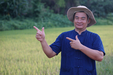 Asian man farmer is at garden, wears hat, blue shirt, point fingers for add text or advertisement, stand at maize garden, feel confident. Concept, Agriculture occupation. Thai farmer lifestyle