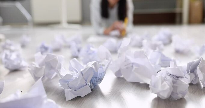 Crumpled paper balls scattering on office floor while annoyed girl ponders in background. Moment suggests creative block and frustration slow motion