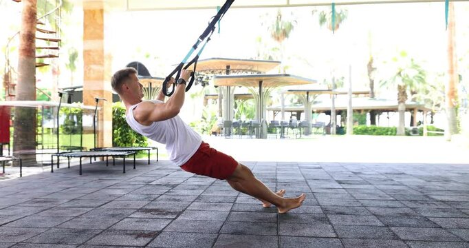 Muscular sportsman performs suspension training exercise outdoor. Man does core strength and physical fitness on public park sports ground slow motion