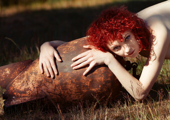 A woman with fiery red hair poses with a weathered, rusty metal object, capturing a blend of beauty...