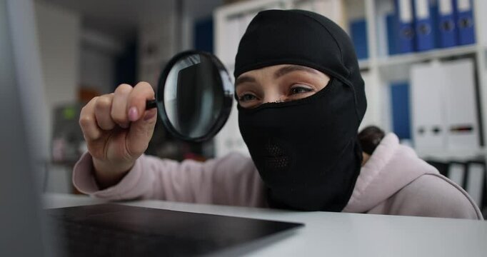 Female hacker in balaclava uses magnifying glass to examine data on laptop screen while sitting at desk in office. Emphasis on theft of personal data - Powered by Adobe