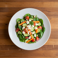 Fresh spinach salad with cherry tomato, cucumber, carrot, and crumbled cheese served on white plate on wooden table, healthy and colorful meal option