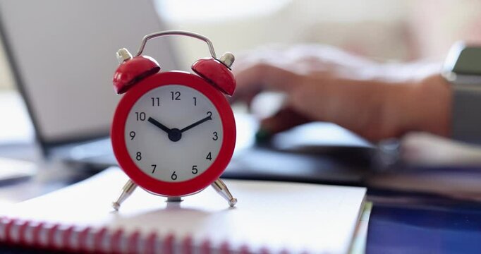 Woman types on laptop keyboard near vibrant red alarm clock on spiral-bound notebook on desk. Focus on productivity and time management in office