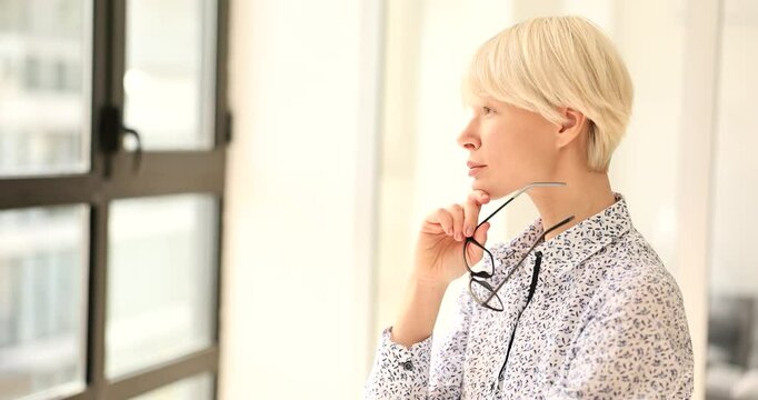 Thoughtful blonde woman stands by large window holding glasses. Experienced businesswoman considers new ways to implement innovative solutions