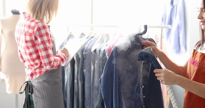 Seamstresses steam clothes on hangers with steam iron at textile factory. Preparation of garments and taking care of finishing touches in dry cleaning