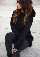 close up young casual brunette girl in black sports hoodie and jeans is sitting outdoor on the big white minimal stairs background and looking away with cute smile, sport lifestyle concept, free space