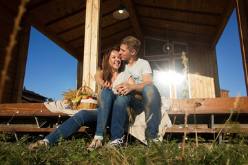 happy pretty couple in jeans and T-shirts is sitting and smiling on the step on the wooden house background on the sunset picnic, young family concept, free space